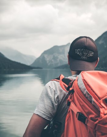man with red hiking backpack facing body of water and mountains at daytime